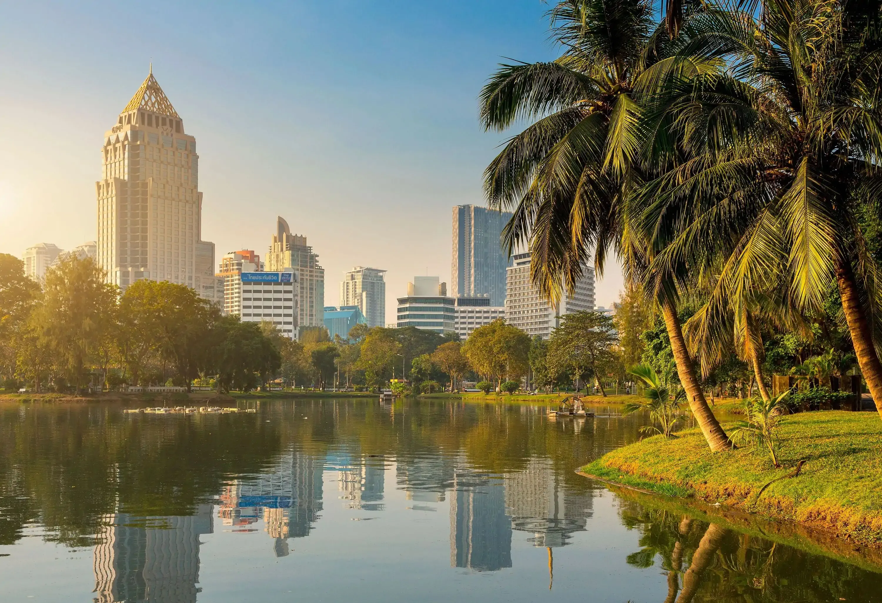 A lush park with an artificial lake lined with tall green trees against the backdrop of an urban cityscape.