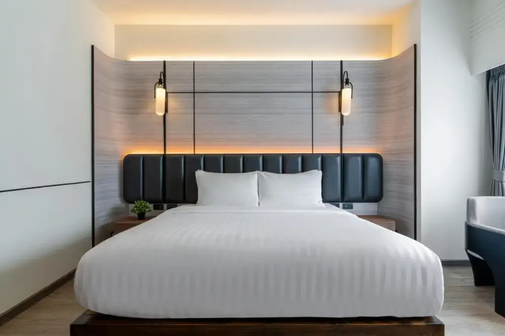 Modern hotel room featuring a large bed with crisp white linens and a black upholstered headboard. Warm ambient lighting and sleek wood paneling create a tranquil atmosphere.