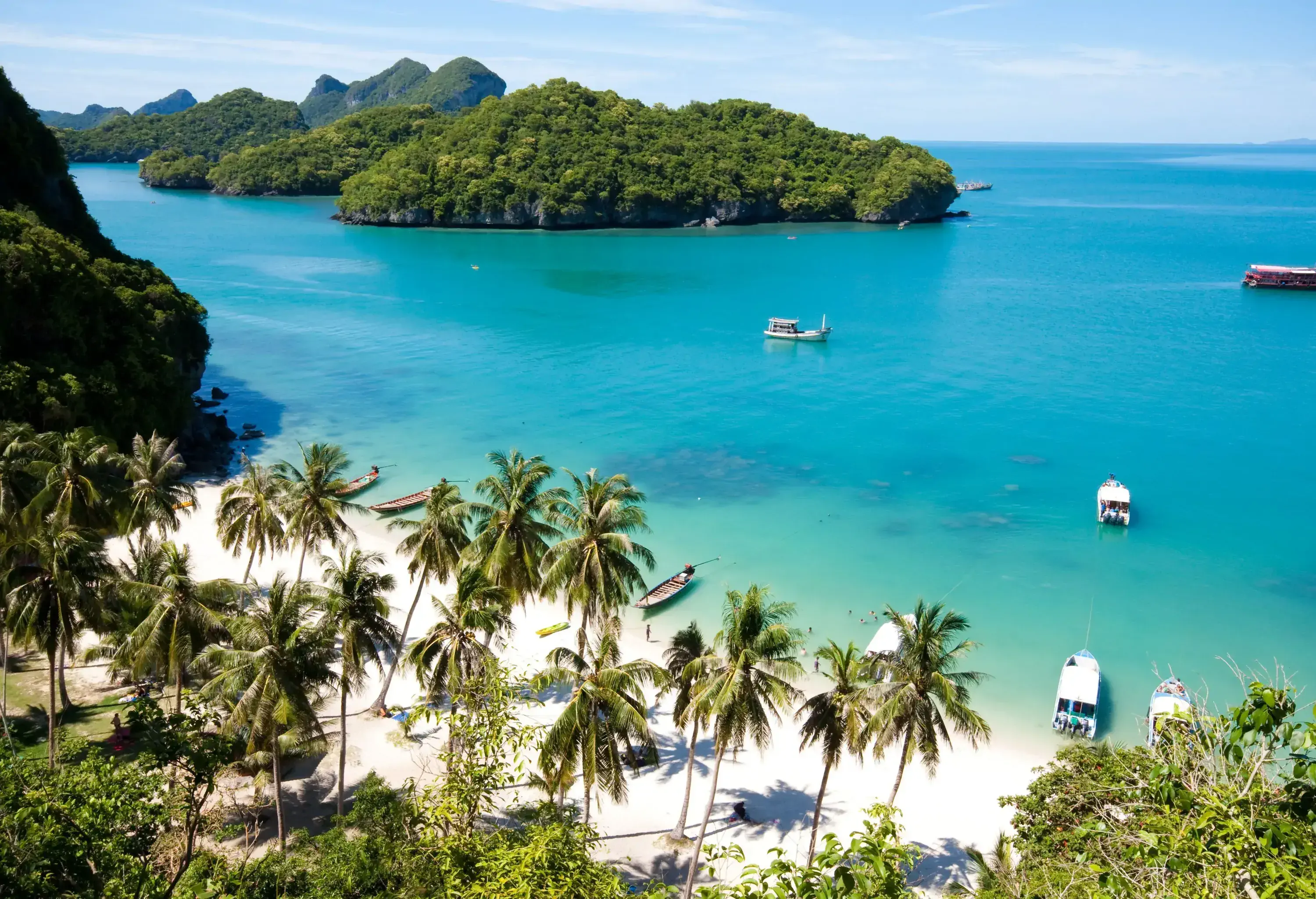 A white sand beach with anchored boats on turquoise waters surrounded by forested islands.