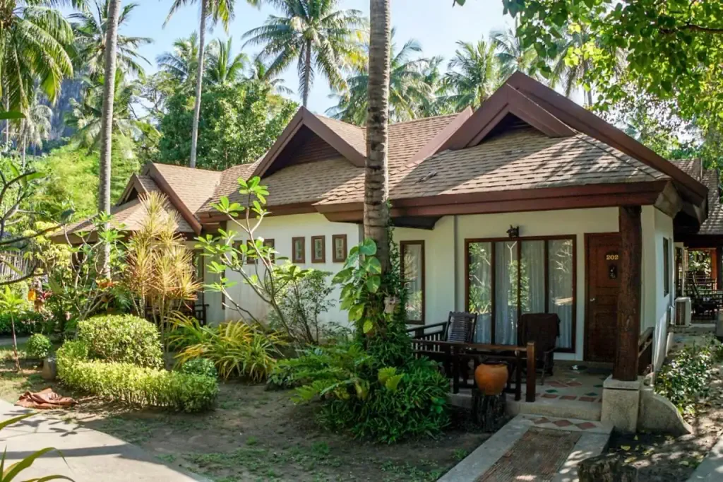A tropical bungalow surrounded by lush greenery and palm trees, featuring a wooden porch with chairs. 
