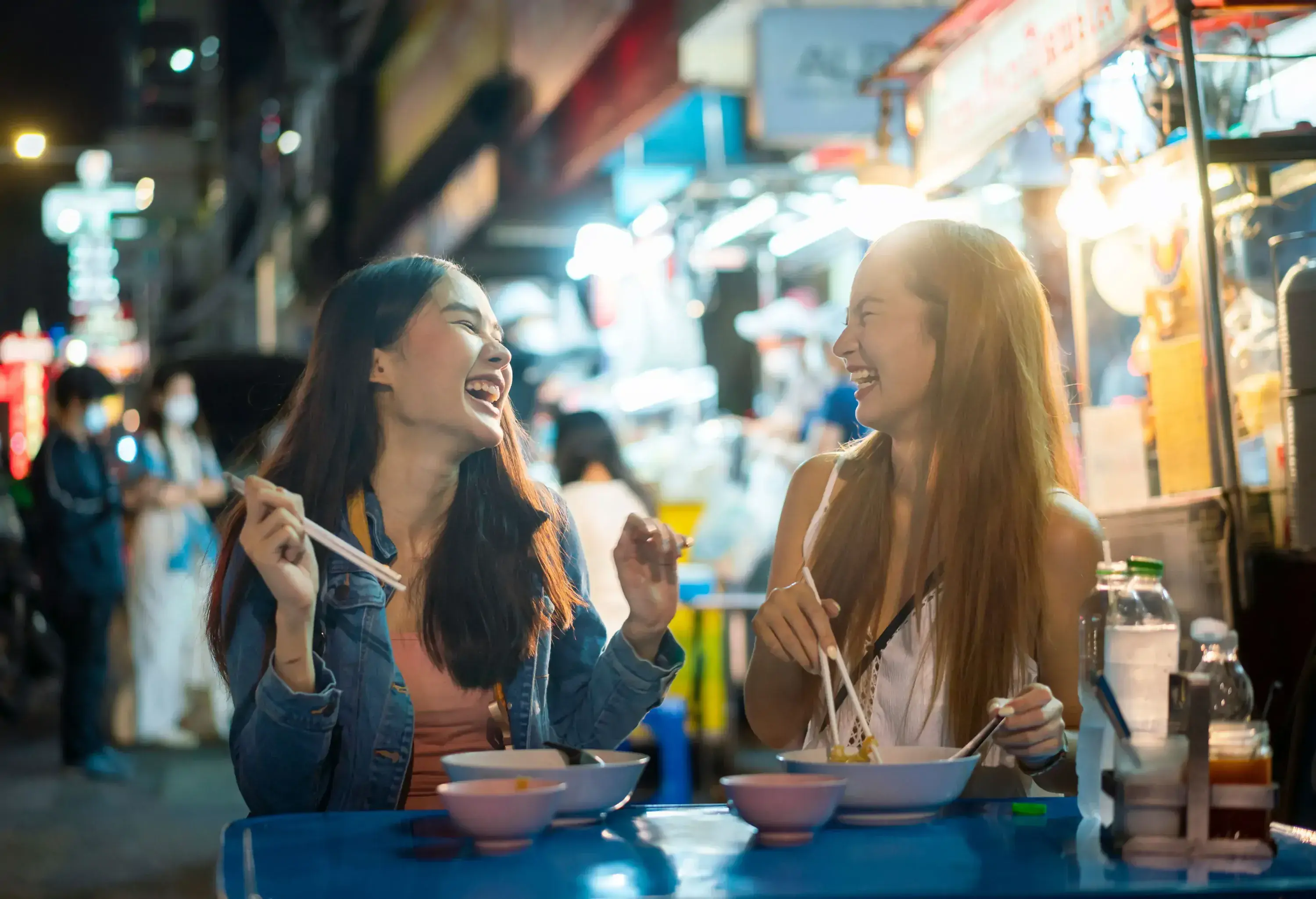 Two happy women sit and dine together on a brightly lit market at night.