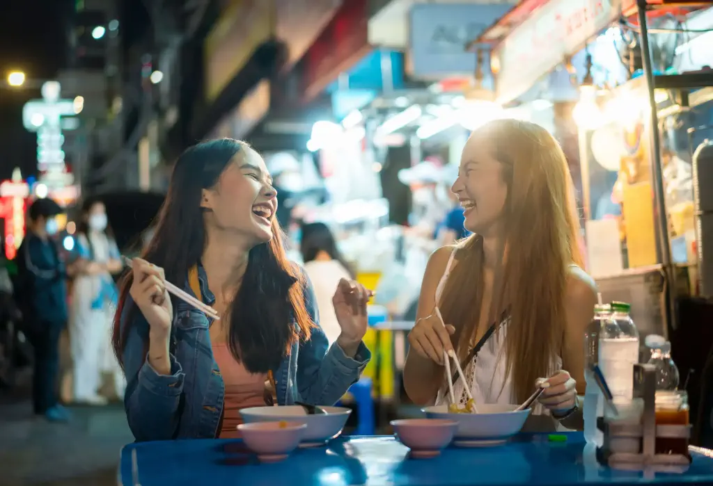 Two happy women sit and dine together on a brightly lit market at night.