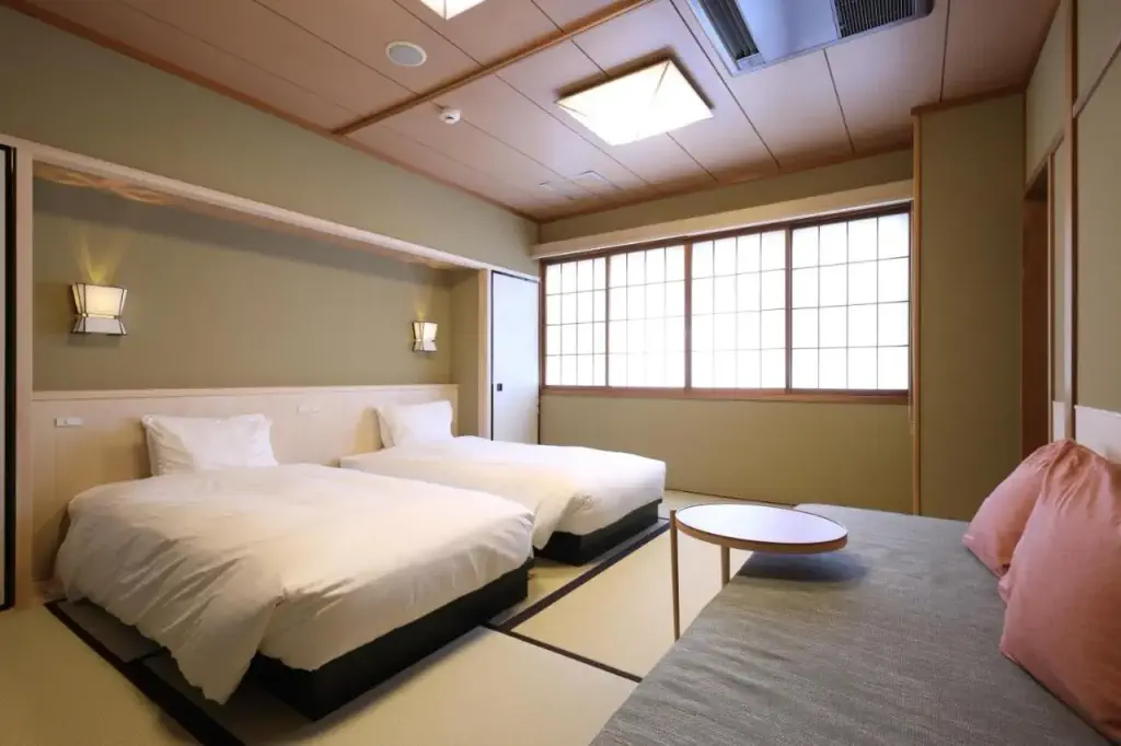 Japanese-style hotel room with two single beds on tatami mats, a sofa with pink cushions, and a round table. Shoji window creates a serene ambiance.