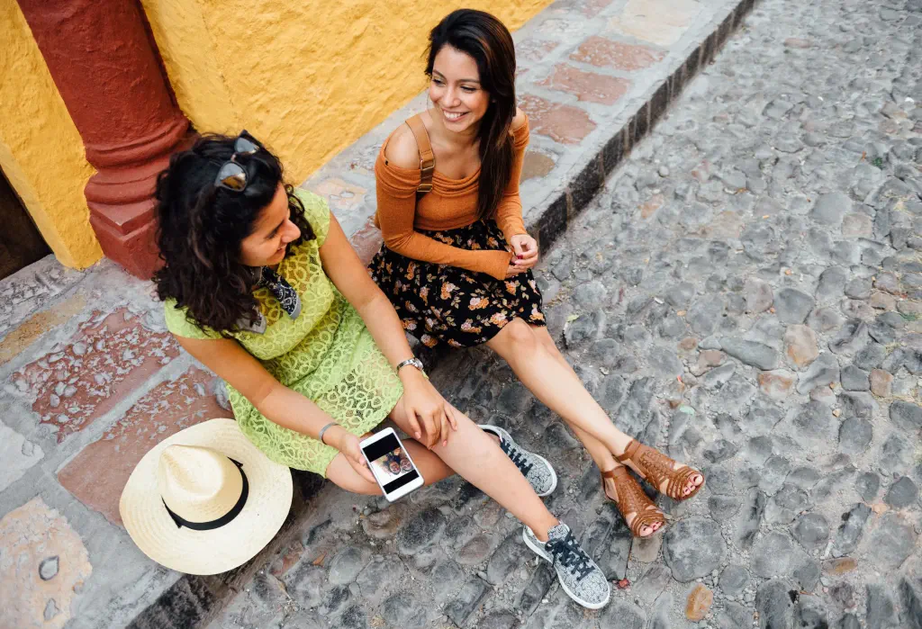 Two happy female friends sit on the sidewalk of a cobbled street.