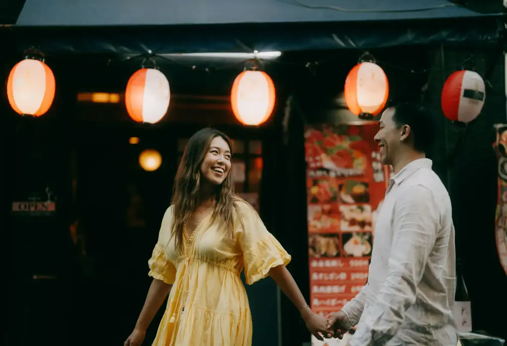 A young couple smiles at each other while holding hands and walking past a building with illuminated paper lanterns.