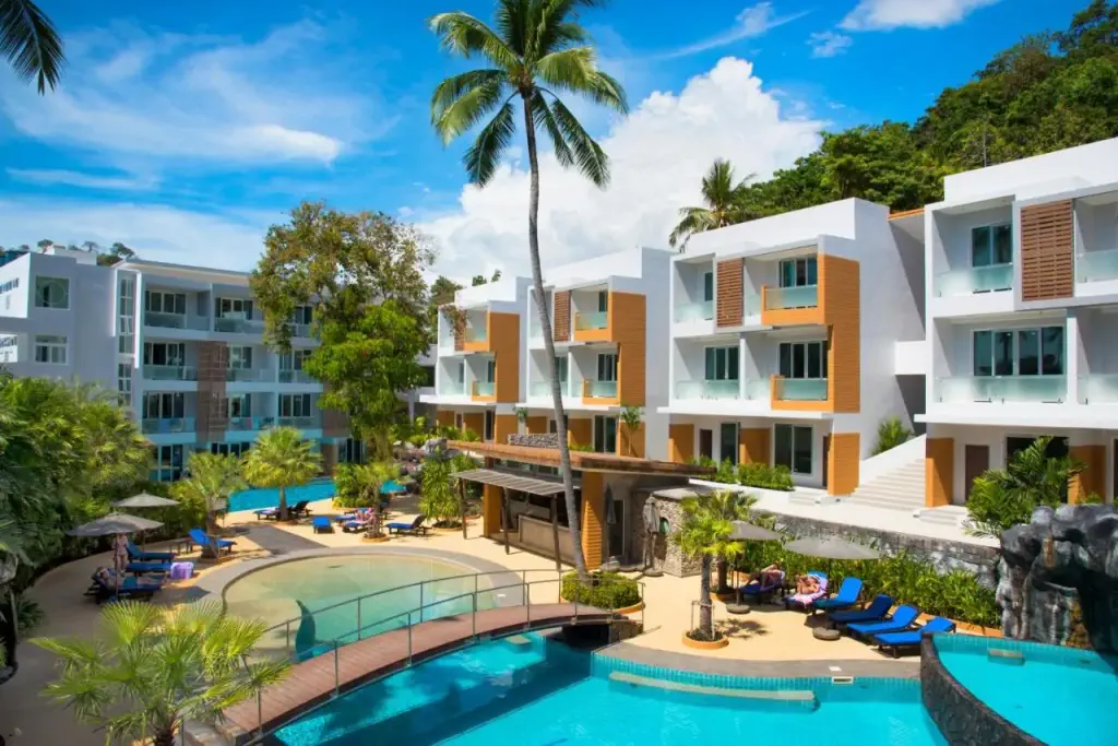 Tropical resort with modern white buildings, palm trees, and a pool under a bright blue sky. Relaxed atmosphere with lounge chairs by the water.