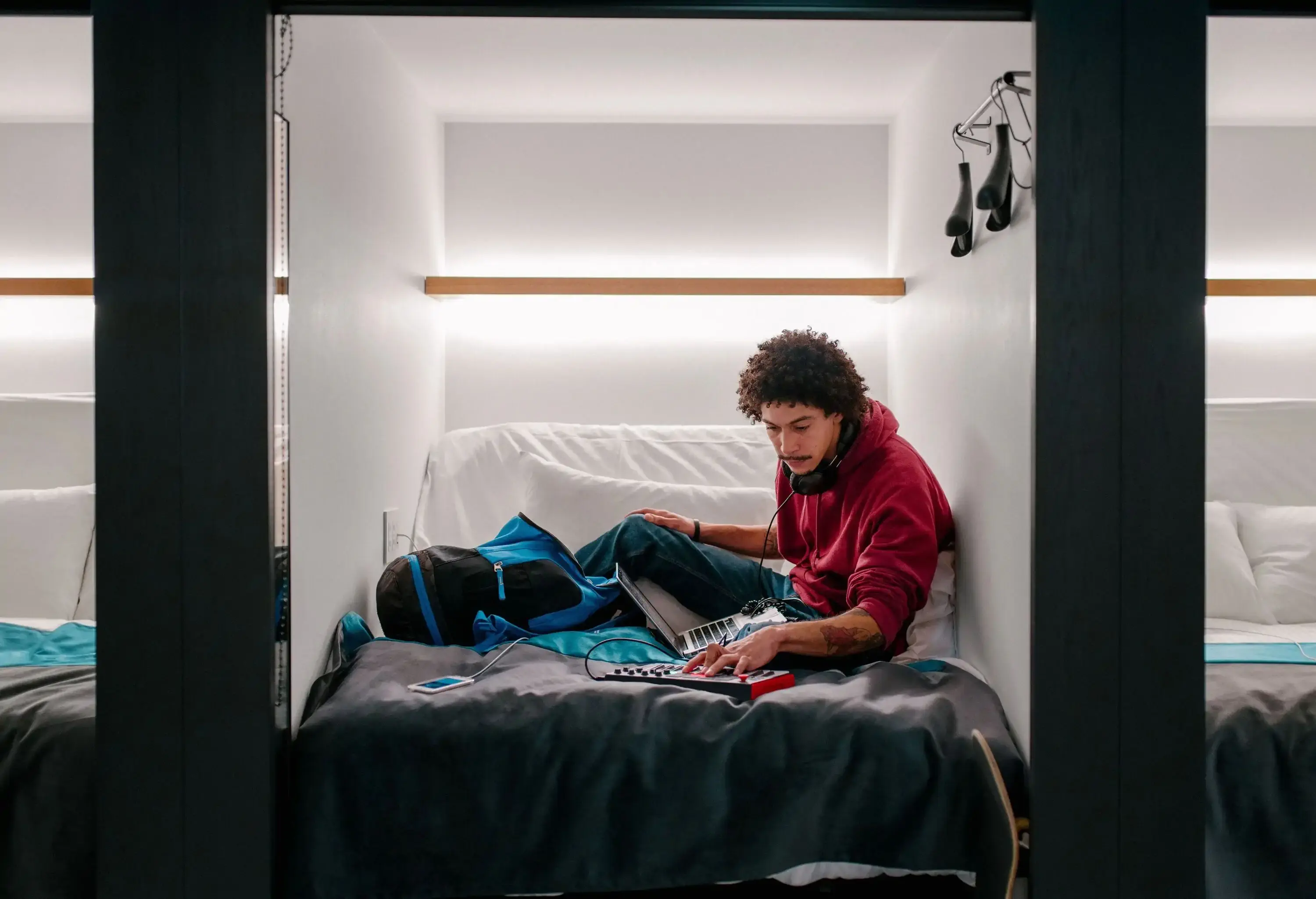 A young adult male relaxes in a capsule hotel, lying down as he engages with audio equipment.