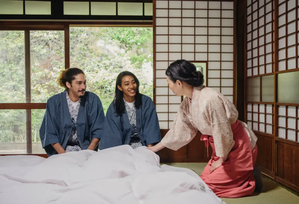 Two young travellers, dressed in kimonos, listen attentively to the hotel owner as she prepares their futon with a warm smile, embodying the values of respect and exceptional service in traditional Japanese hospitality.