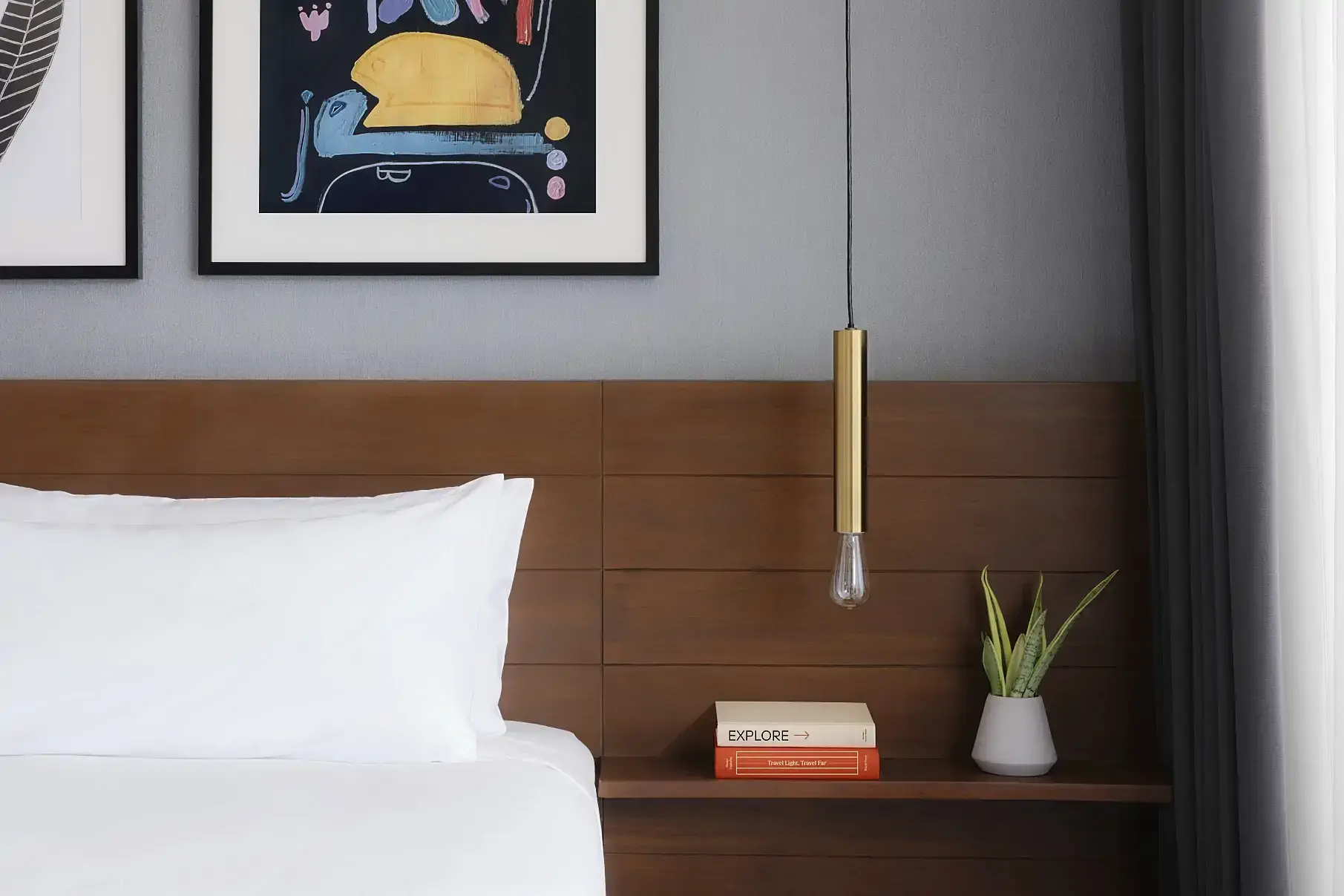 A modern bedroom with a wooden headboard, white bed linens, and two framed artworks above. One artwork is abstract and colorful, and the other features a black leaf design on a white background. A pendant light hangs beside a small plant and books on the headboard, next to sheer curtains. The tone is calm and minimalist.
