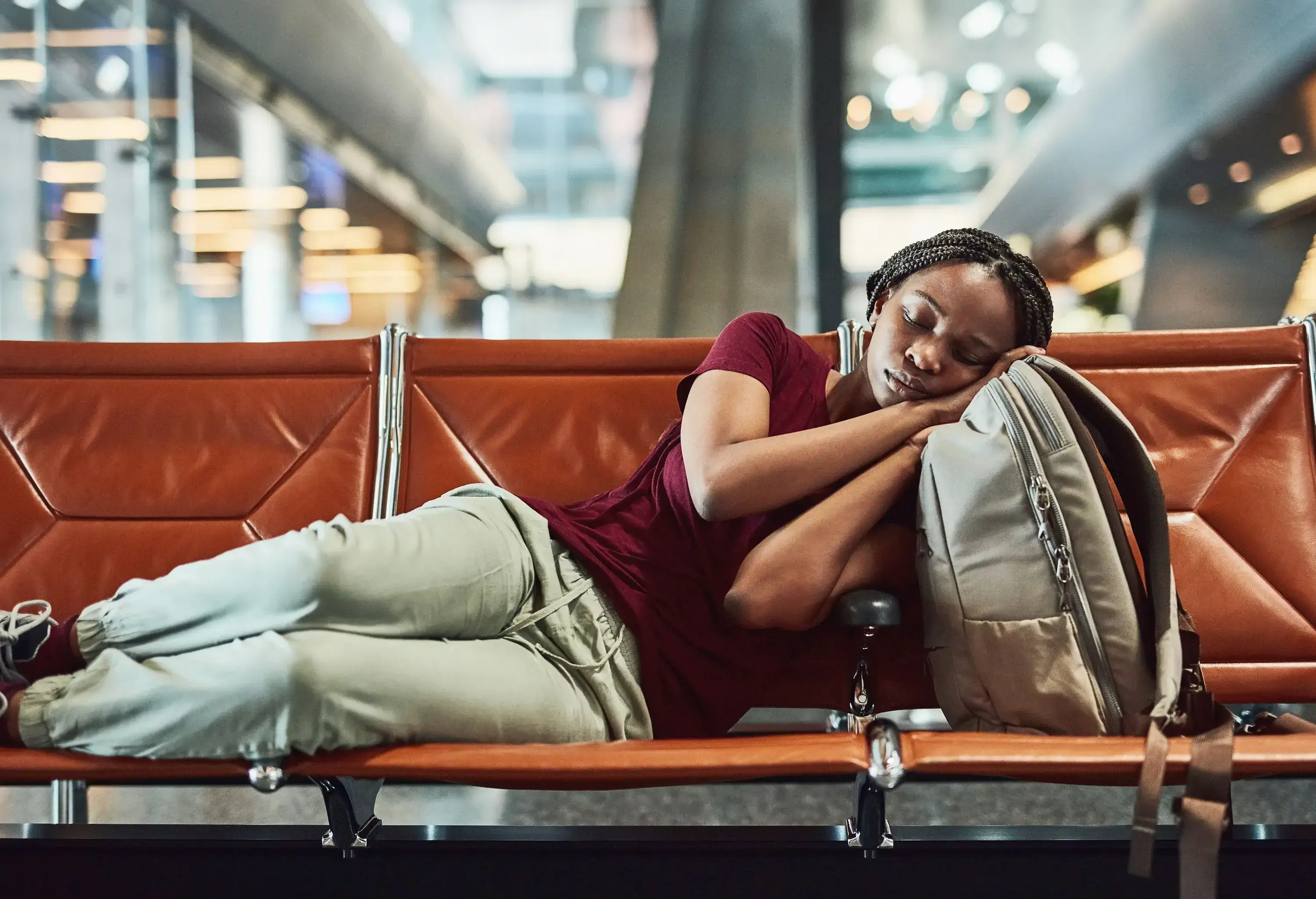 A woman sleeping on an airport bench, resting her head on a backpack.