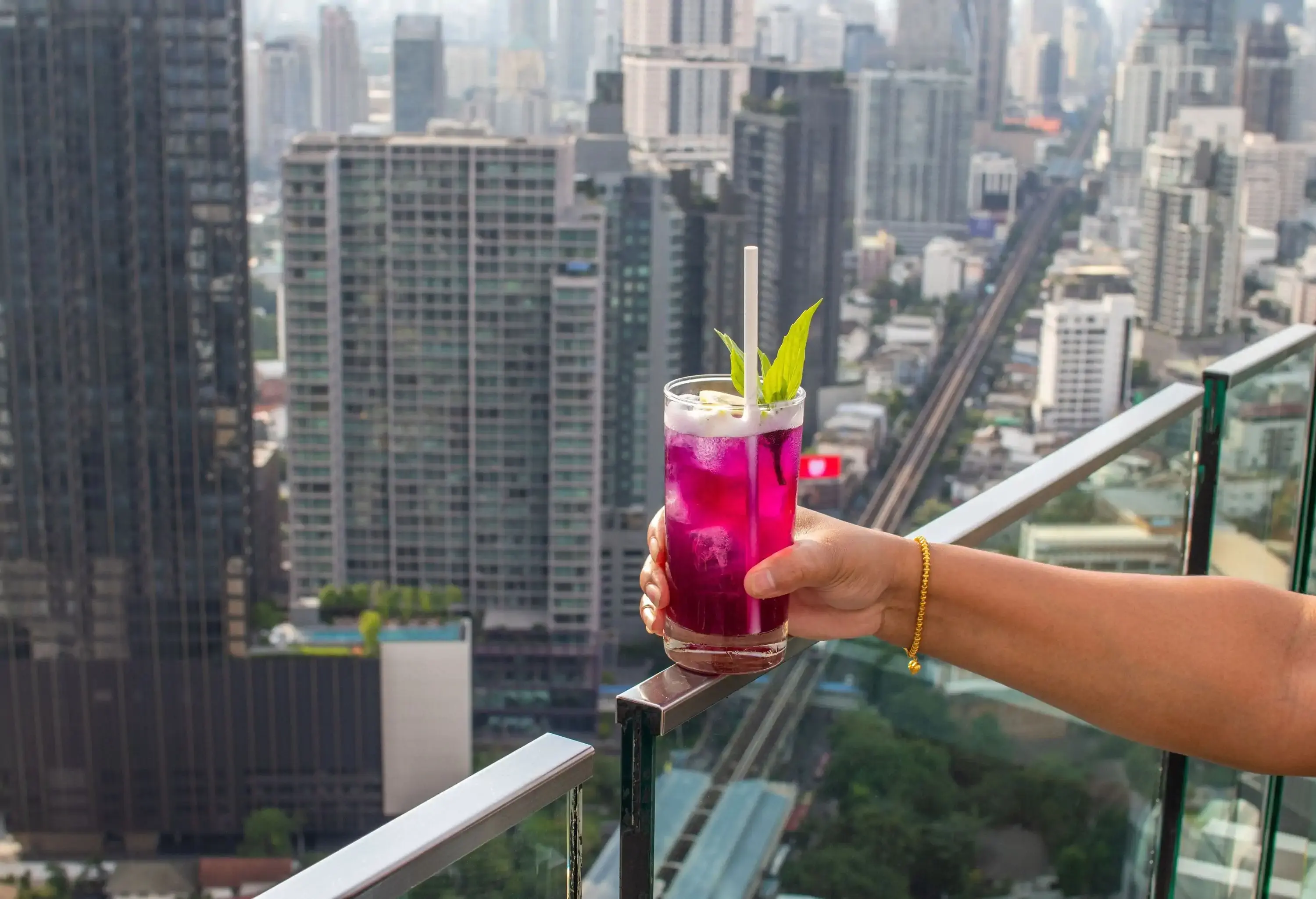 A hand holding a cocktail over the glass barrier of a rooftop bar.