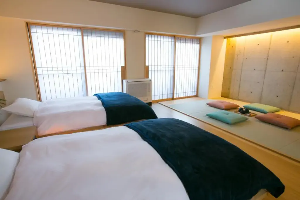 A serene bedroom with two beds and dark green blankets facing a Japanese tatami mat area. Soft lighting and sliding shoji screens create a tranquil ambiance.