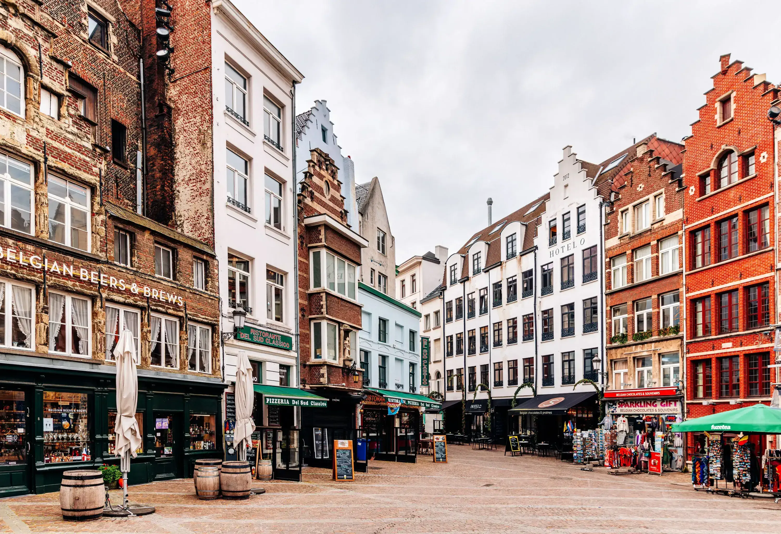 A picturesque town square lined with old brick buildings, housing ground-floor shops and restaurants under a cloudy sky.