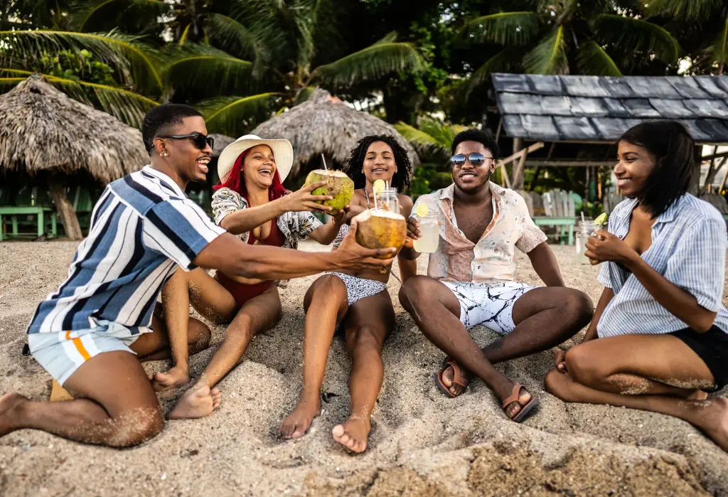 Five cheerful individuals sitting on a sandy beach, sharing a laugh and raising their coconut and glass drinks in a toast.