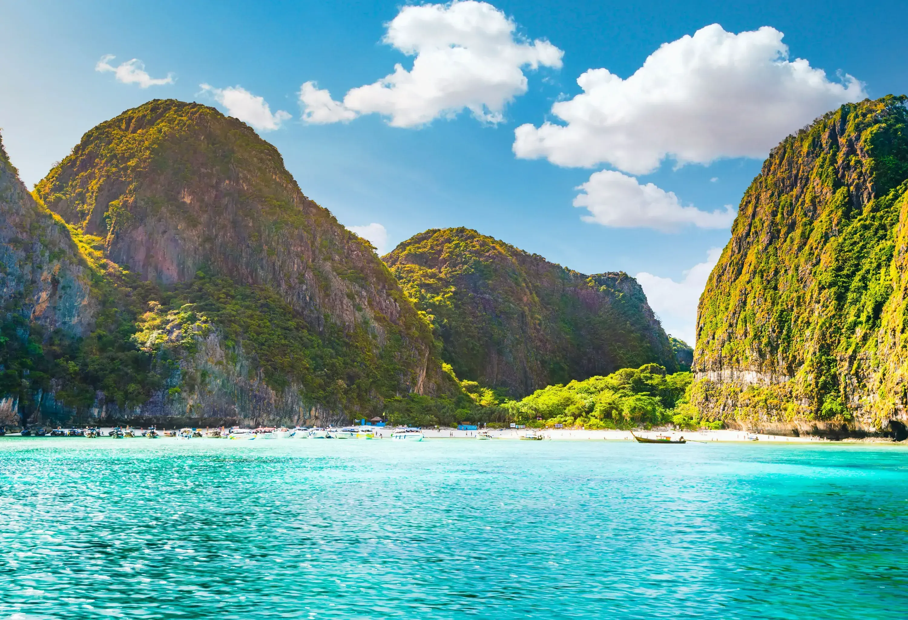 Several boats line up along the white sand turquoise beach bounded by lush hills.