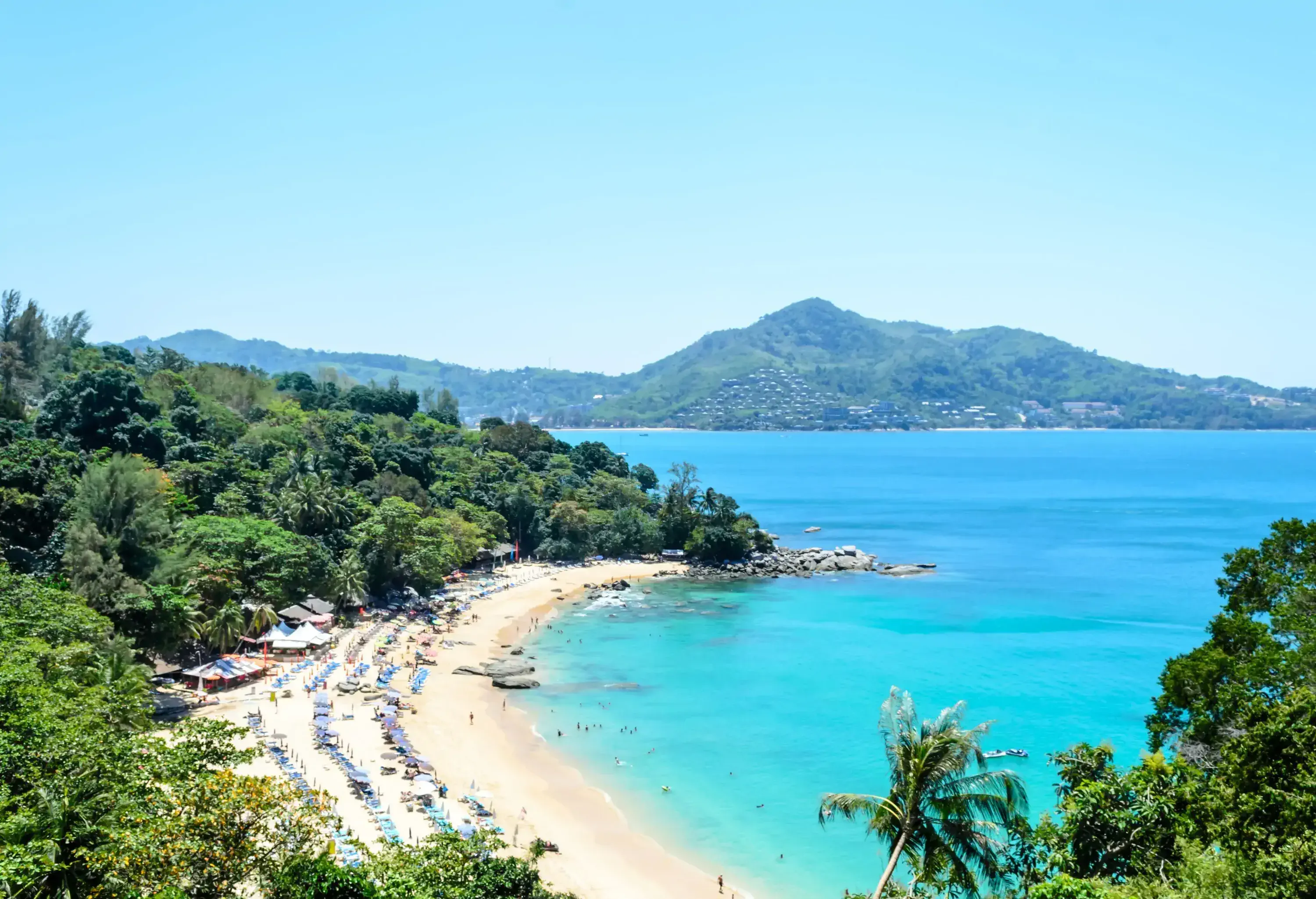 A crowded beach with sunbeds and umbrellas along a forested hill with views of a large bay.
