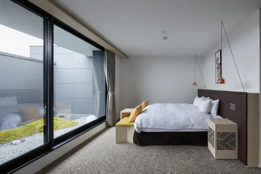 Modern bedroom with a large window, minimalist decor, and neutral tones. A cozy double bed has white linens, yellow pillows, and hanging copper lamps.