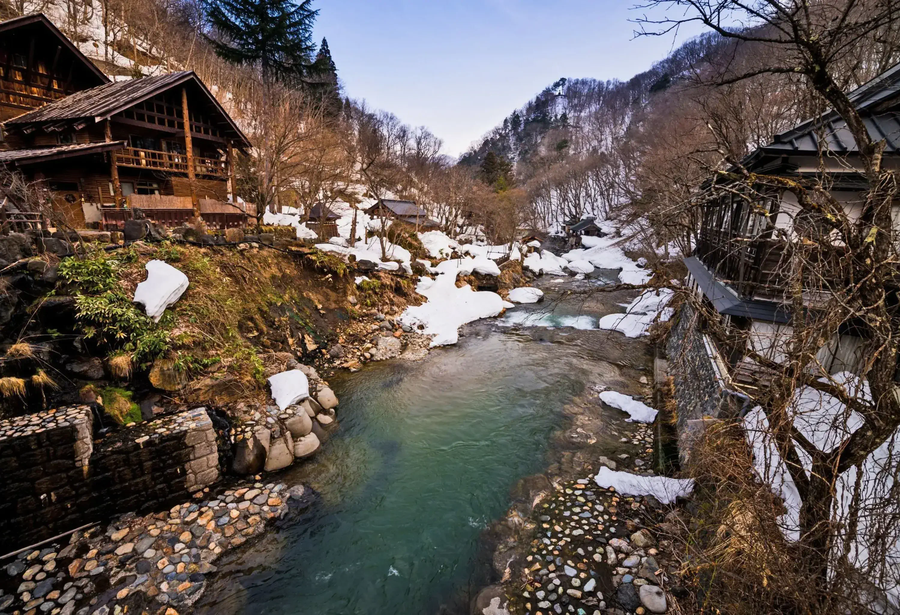 A small river across mountain cabins amongst bare trees and heavy snow.
