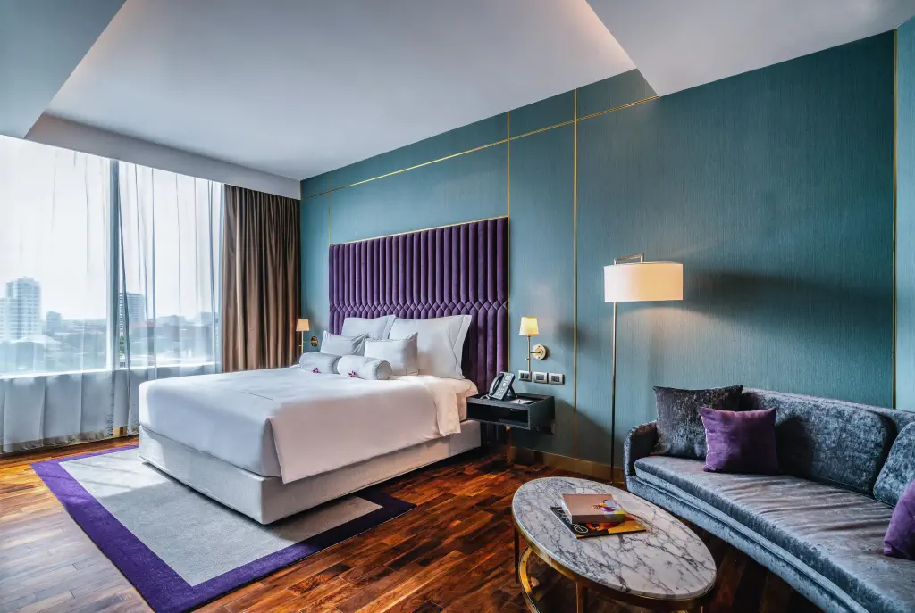 Luxurious hotel room with a large bed, purple velvet headboard, teal walls, and elegant decor. 