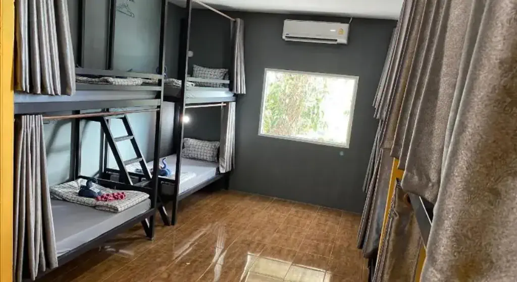 Room with four bunk beds, each with curtains, pillows, and blankets. A window lets in sunlight, and an air conditioner is above. The floor is tiled.
