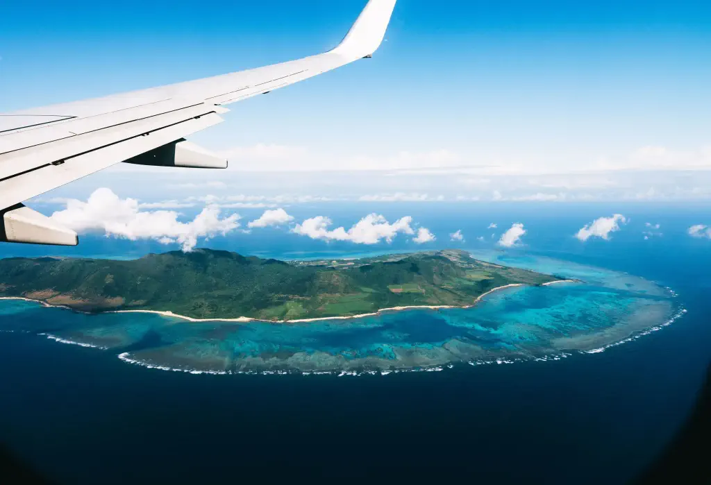 An aeroplane flying over a tropical island surrounded by the turquoise sea under the cloudy blue sky.