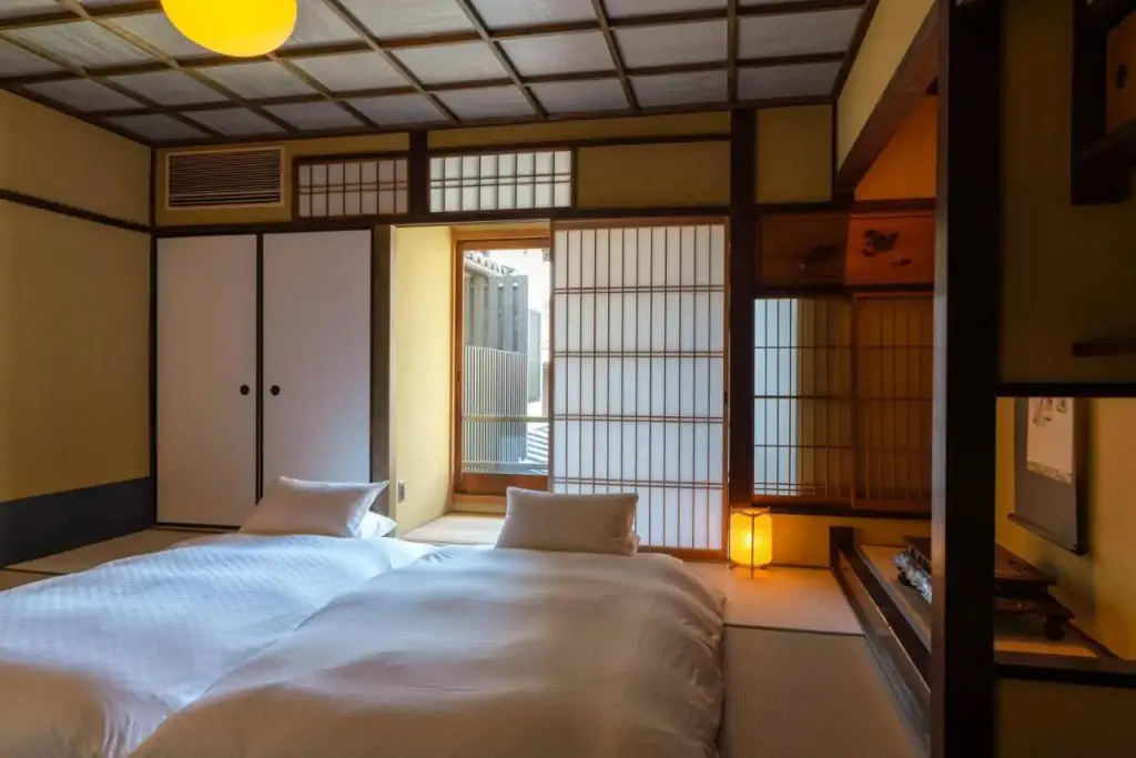 Japanese-style room with two futons, tatami floors, and shoji screens. Soft lighting from a paper lantern adds warmth and a serene atmosphere.