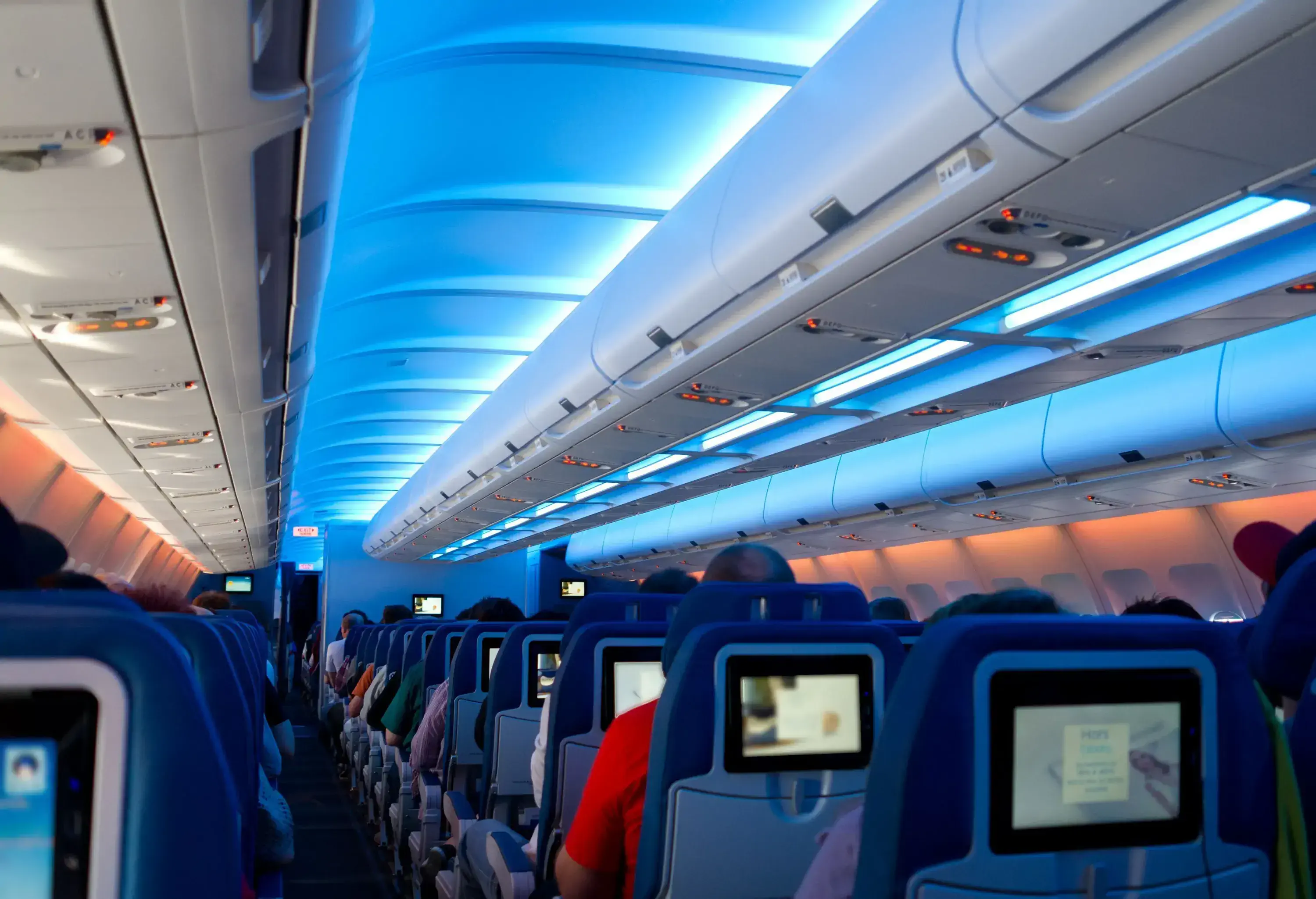 Passengers are seated inside an aircraft with rows of blue chairs featuring small screens, accompanied by soft blue lighting emanating from the ceiling.