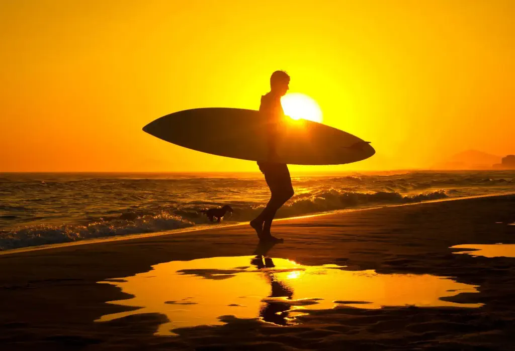 A silhouetted surfer strolls along the beach at sunset, their figure gracefully accentuated against the vibrant hues of the setting sun.