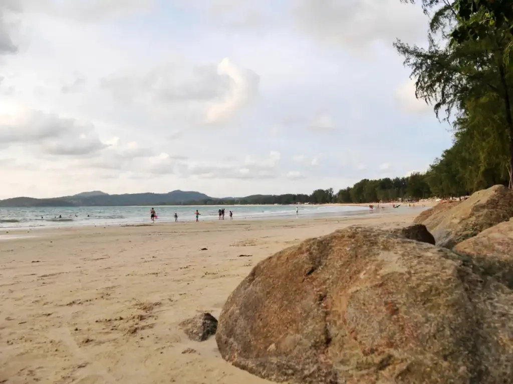 A beach with boulders and large stretch of sand at sunset, people playing in the water