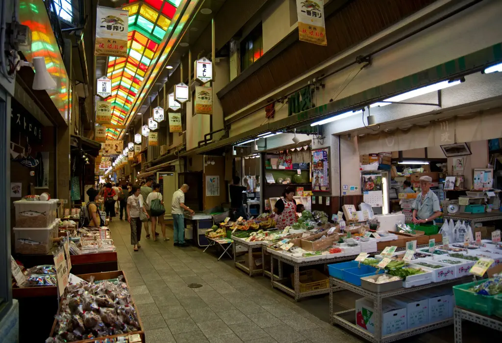 A bustling lane in a vibrant market, illuminated by skylights, is filled with people navigating through a clean environment, showcasing a diverse array of merchandise in lively stalls.