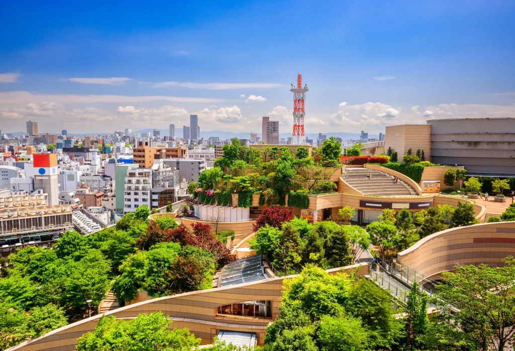 Aerial view of a city featuring terraced green spaces in the foreground, leading to a prominent tower and high-rises against a clear blue sky.