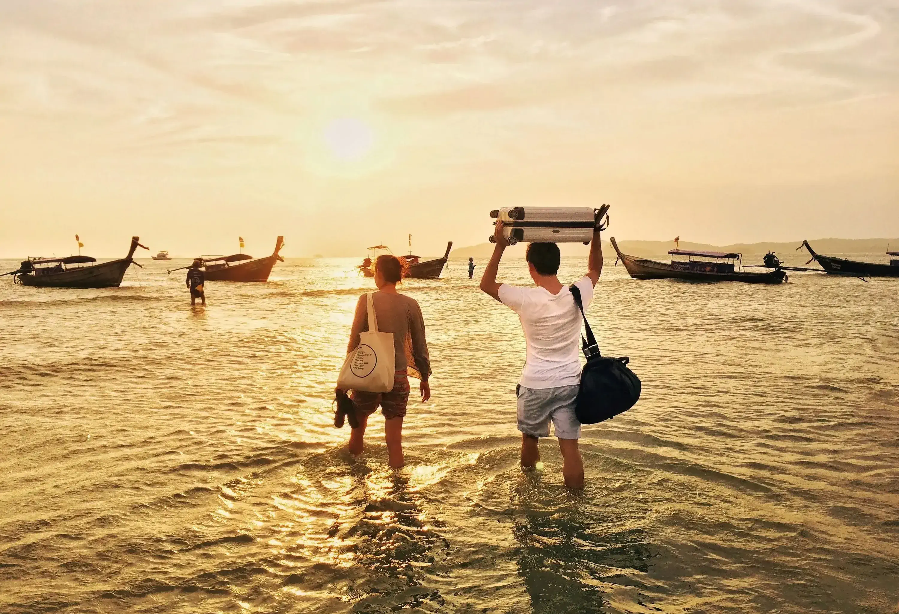 A couple carrying their bags and luggage walk into the sunlit water heading towards the docked boats.