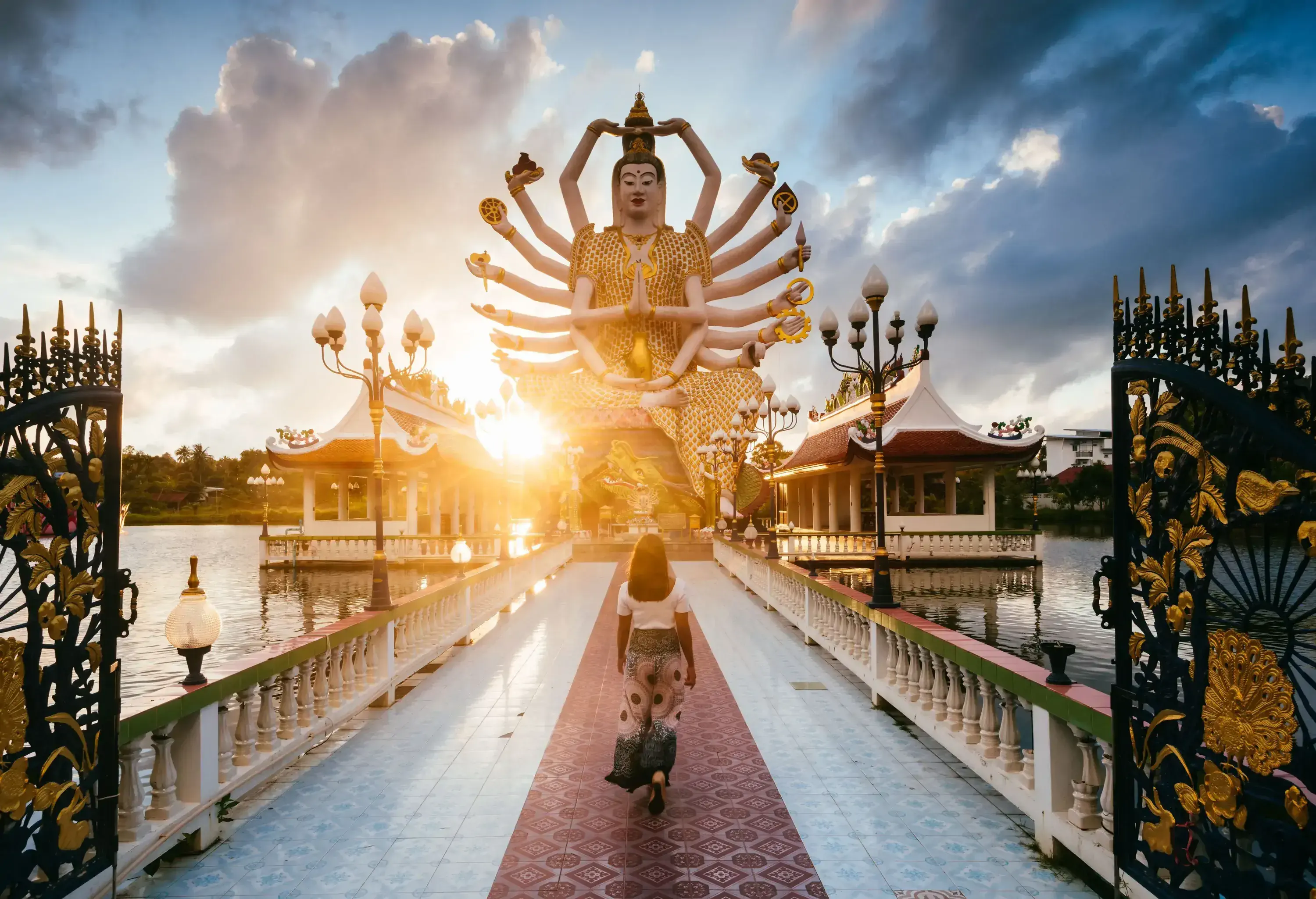 A woman walks across a bridge that leads to a temple with a big statue of a deity with multiple arms.