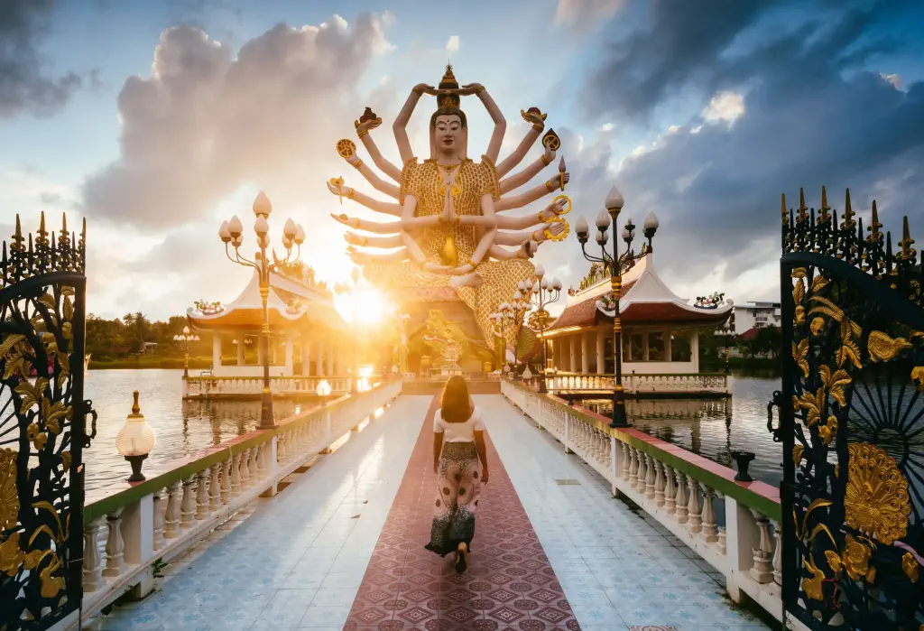 A woman walks across a bridge that leads to a temple with a big statue of a deity with multiple arms.