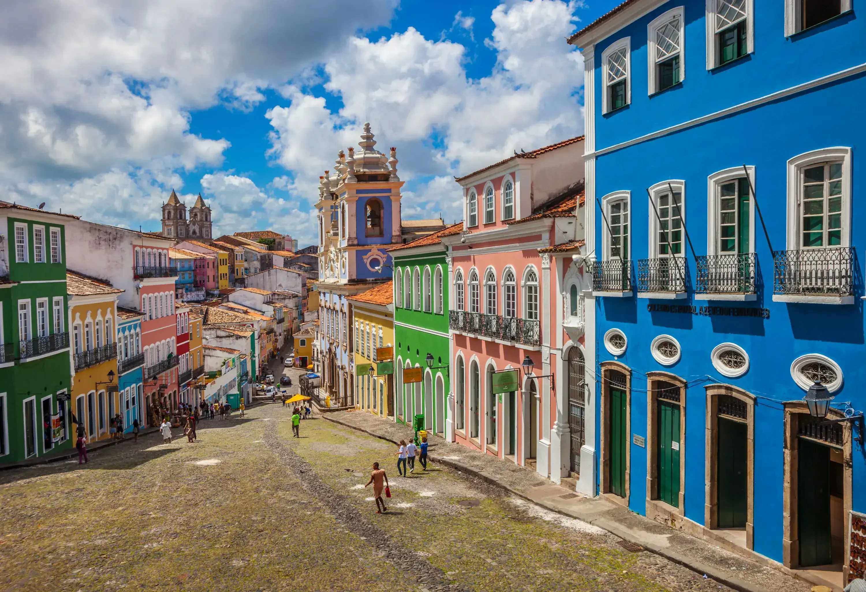 People stroll across a downhill street lined with colourful adjacent buildings.