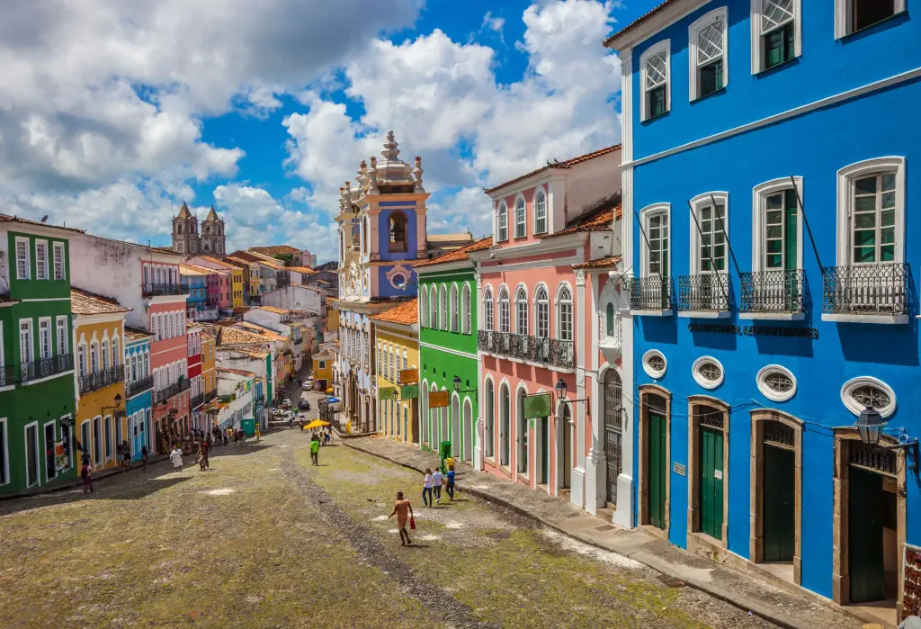 People stroll across a downhill street lined with colourful adjacent buildings.