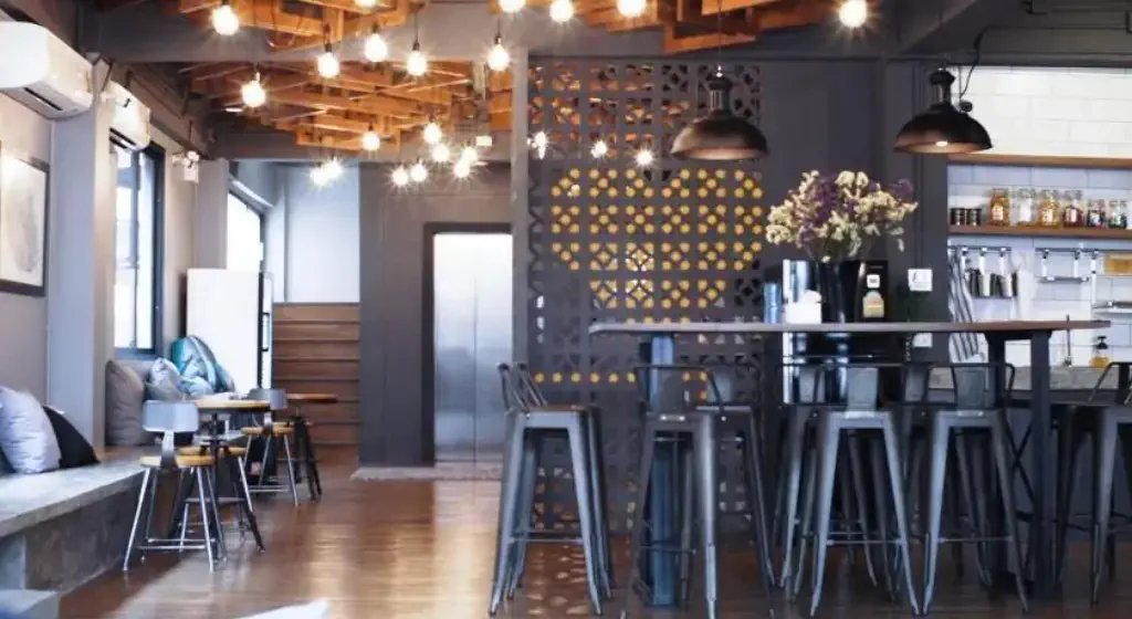 Cozy cafe interior with wooden floors, modern metal chairs, and soft lighting. Decor includes hanging bulbs, floral centerpiece, and a decorative wall partition.