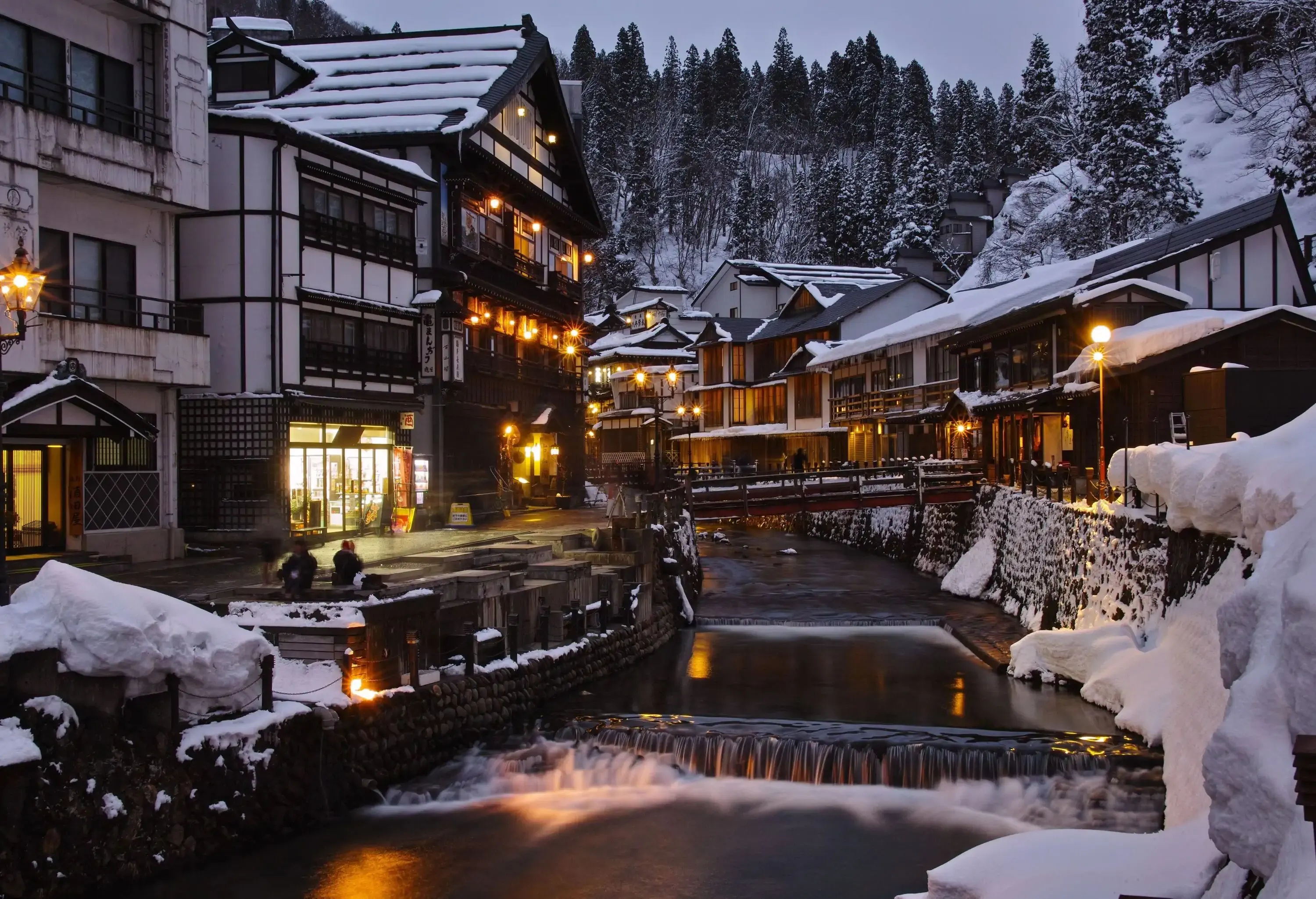 Japanese-style hotels and buildings on each side of a river backed by tall trees and snow-covered mountains. 