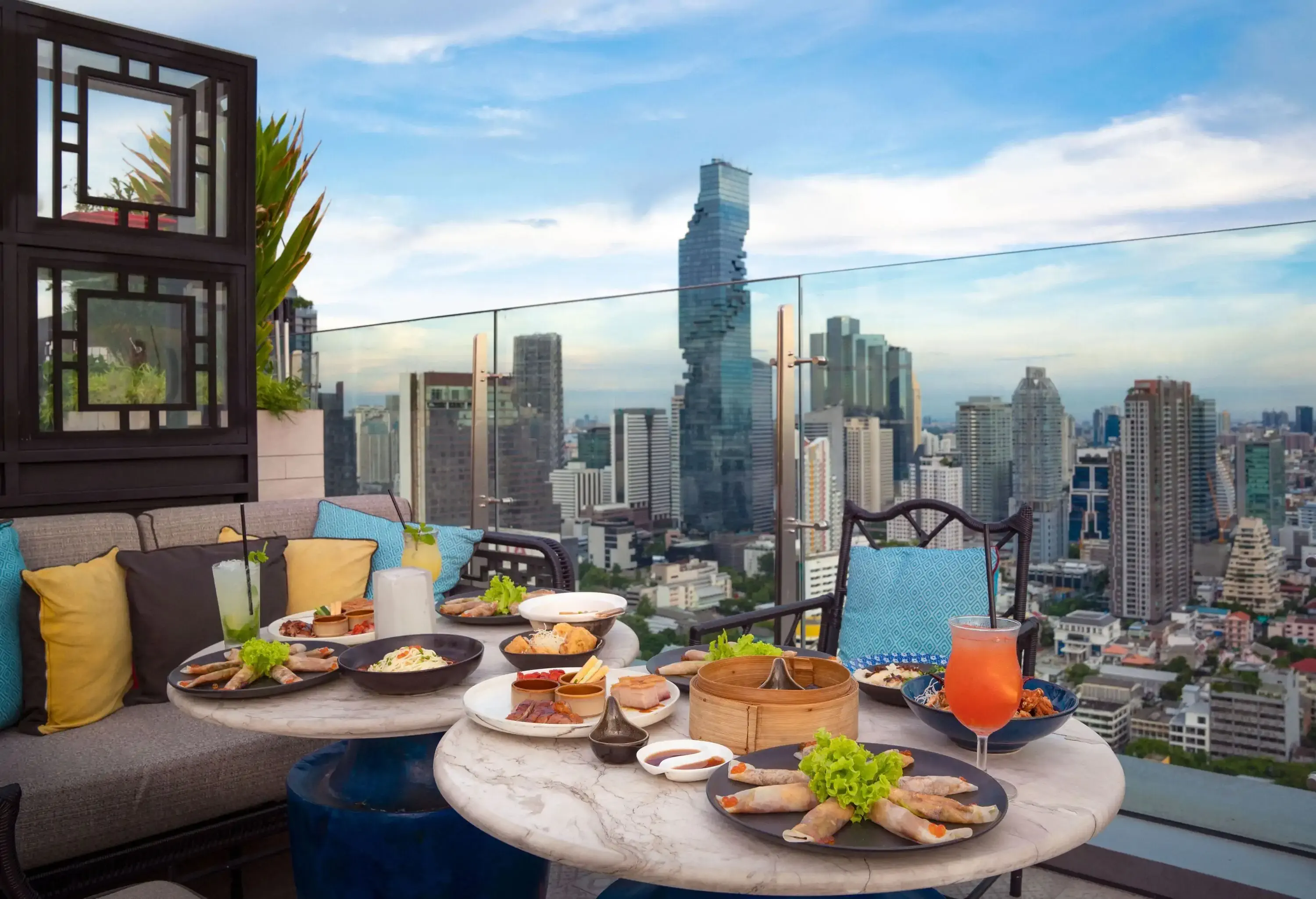 A rooftop dining area with a delectable array of food and beverages on the table and a view of the city skyline.