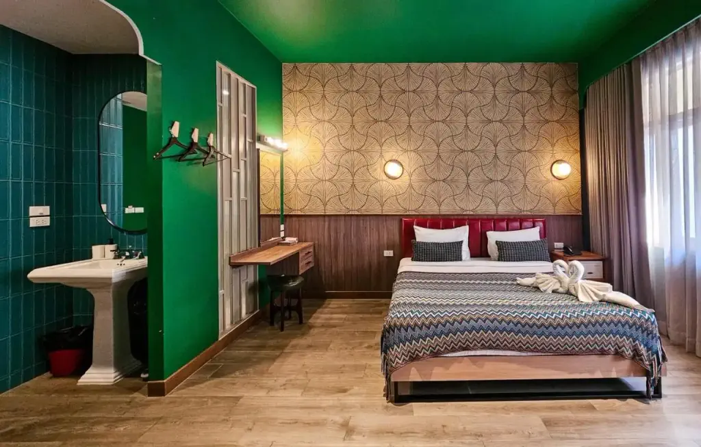 A modern hotel room with a green and brown color scheme. It features a king-sized bed with patterned bedding, two bedside lamps, and a towel swan. The room includes a vanity desk, a green-tiled sink area, and a window with sheer curtains. 