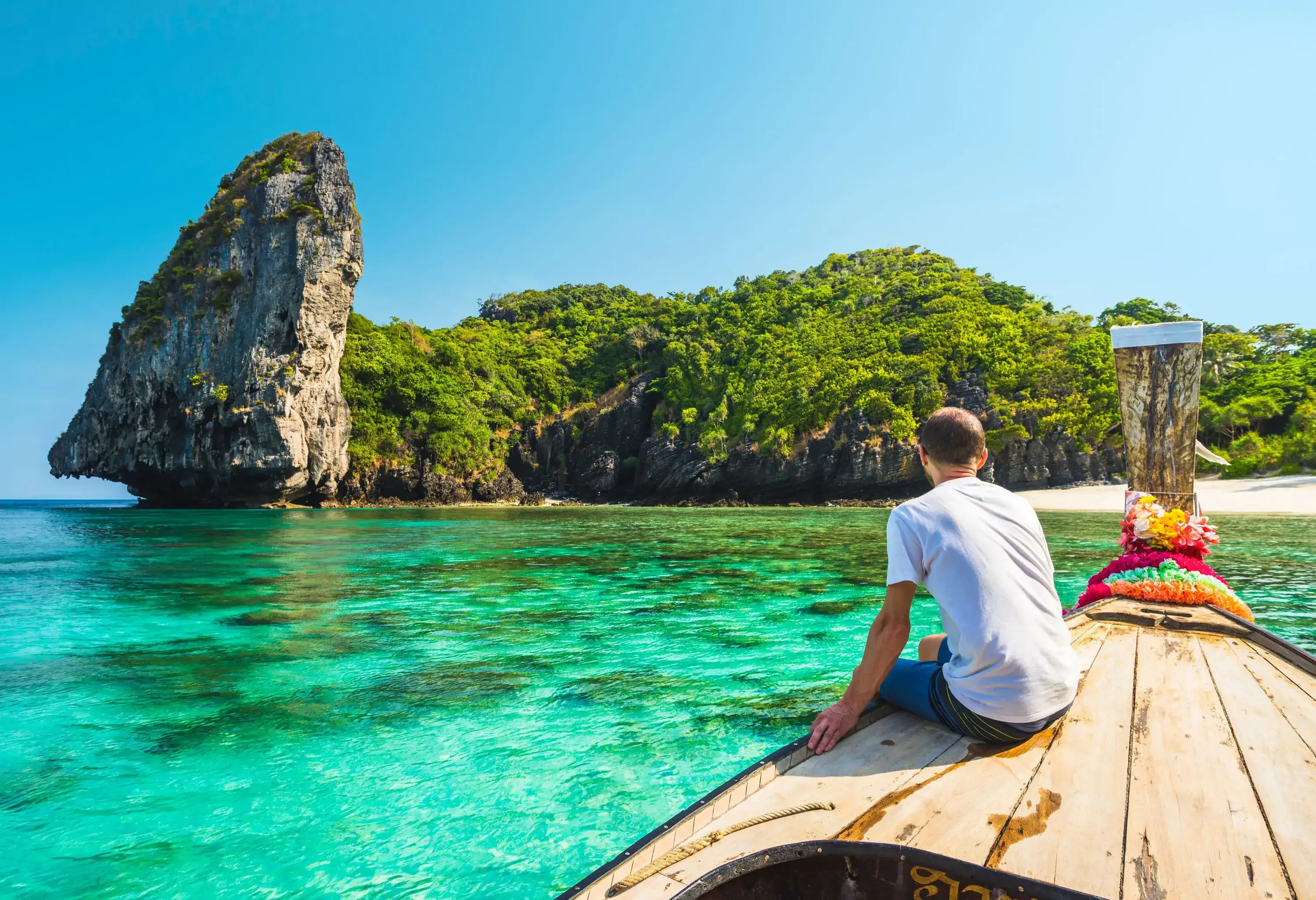 A guy sits aboard a floating long-tail boat, looking out towards the wooded cliff along the beach.