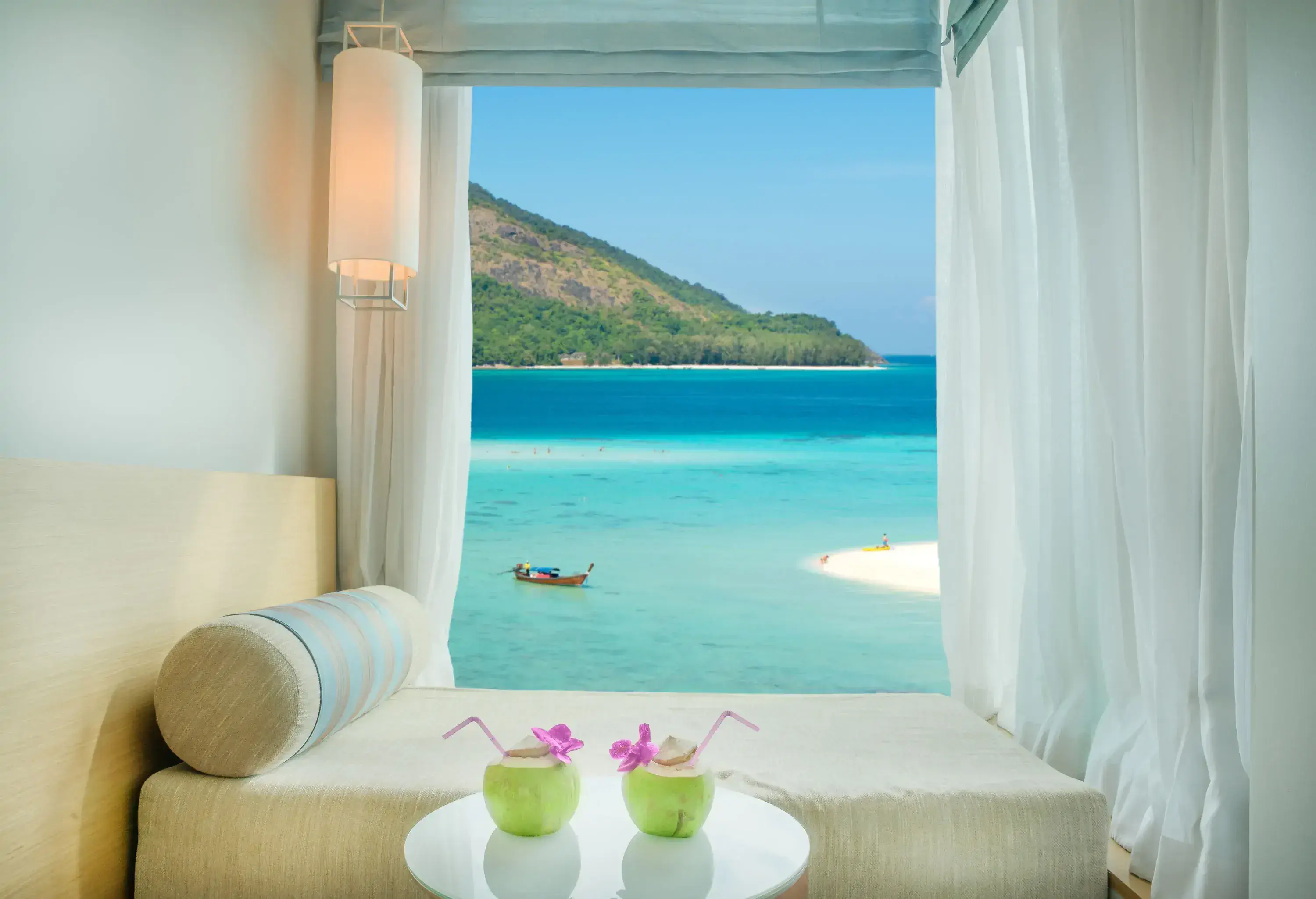 A hotel room with a bed and a table with two coconut drinks and a big window overlooking a boat floating in pristine waters.