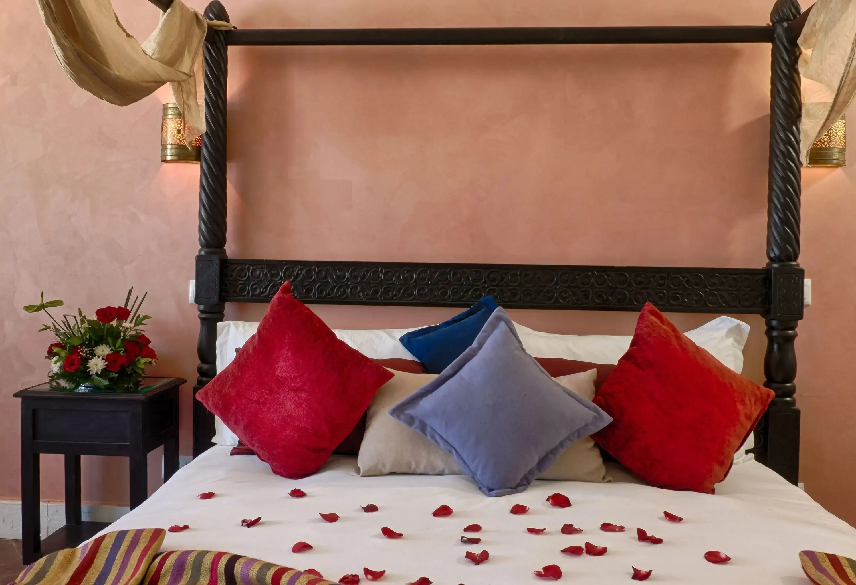 A black canopy bed finished with a white-linen mattress decorated with colourful throw pillows and red rose petals.
