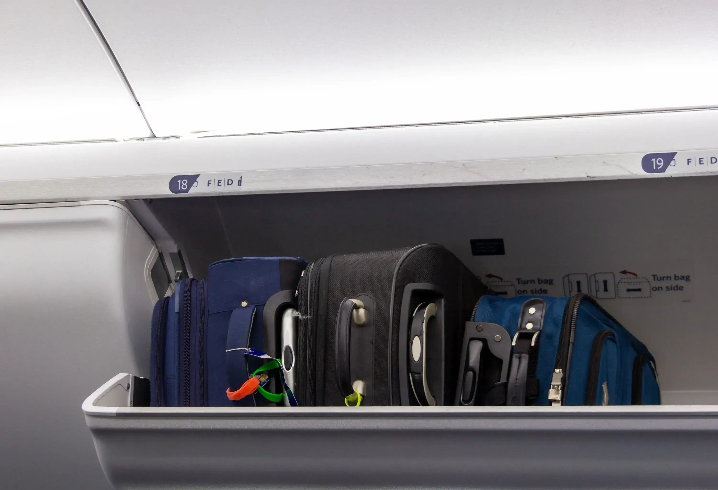 An open overhead compartment on an airplane with three suitcases inside.
