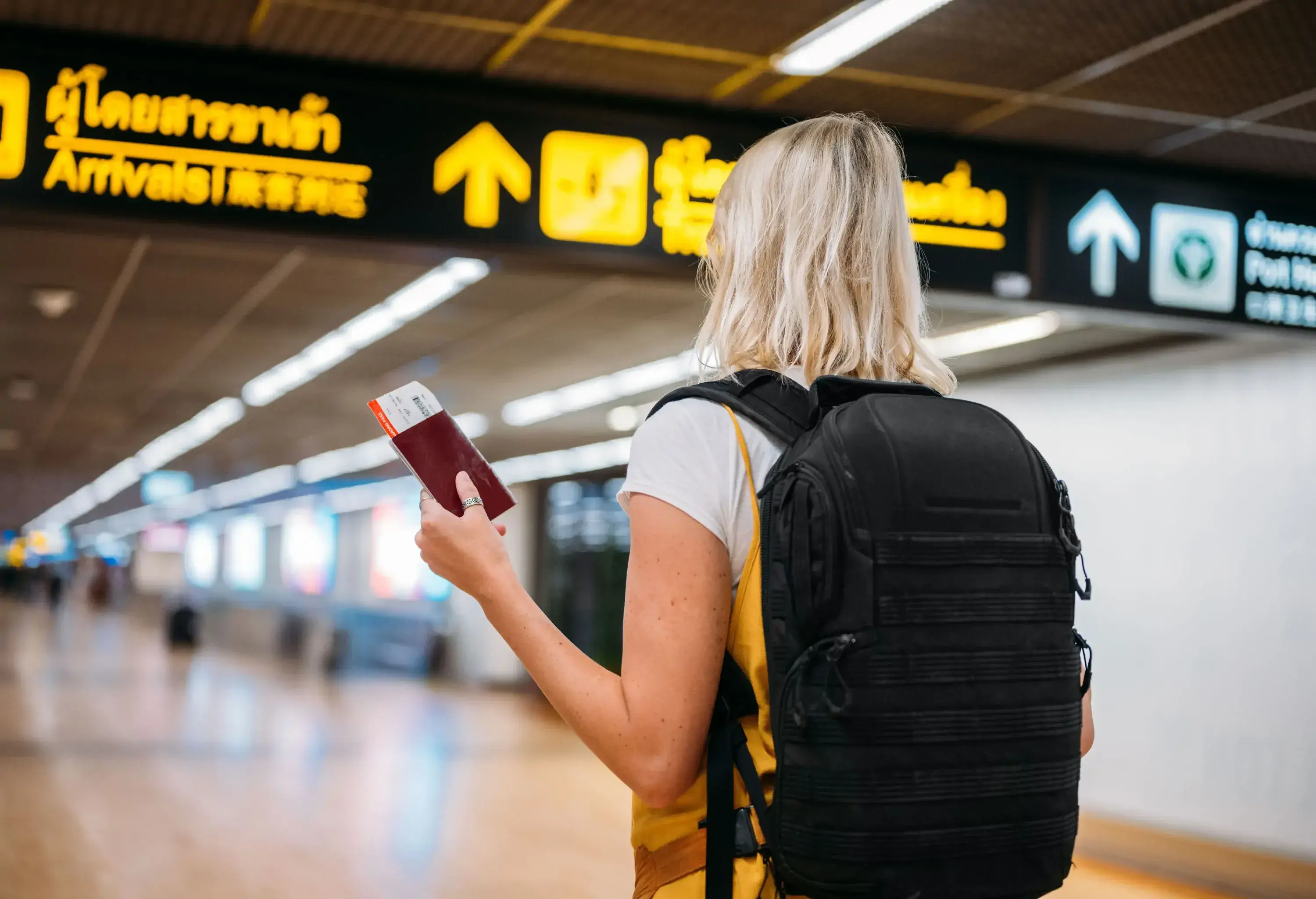 A woman with a black backpack holding her passport and looking at the airport's overhead signs.