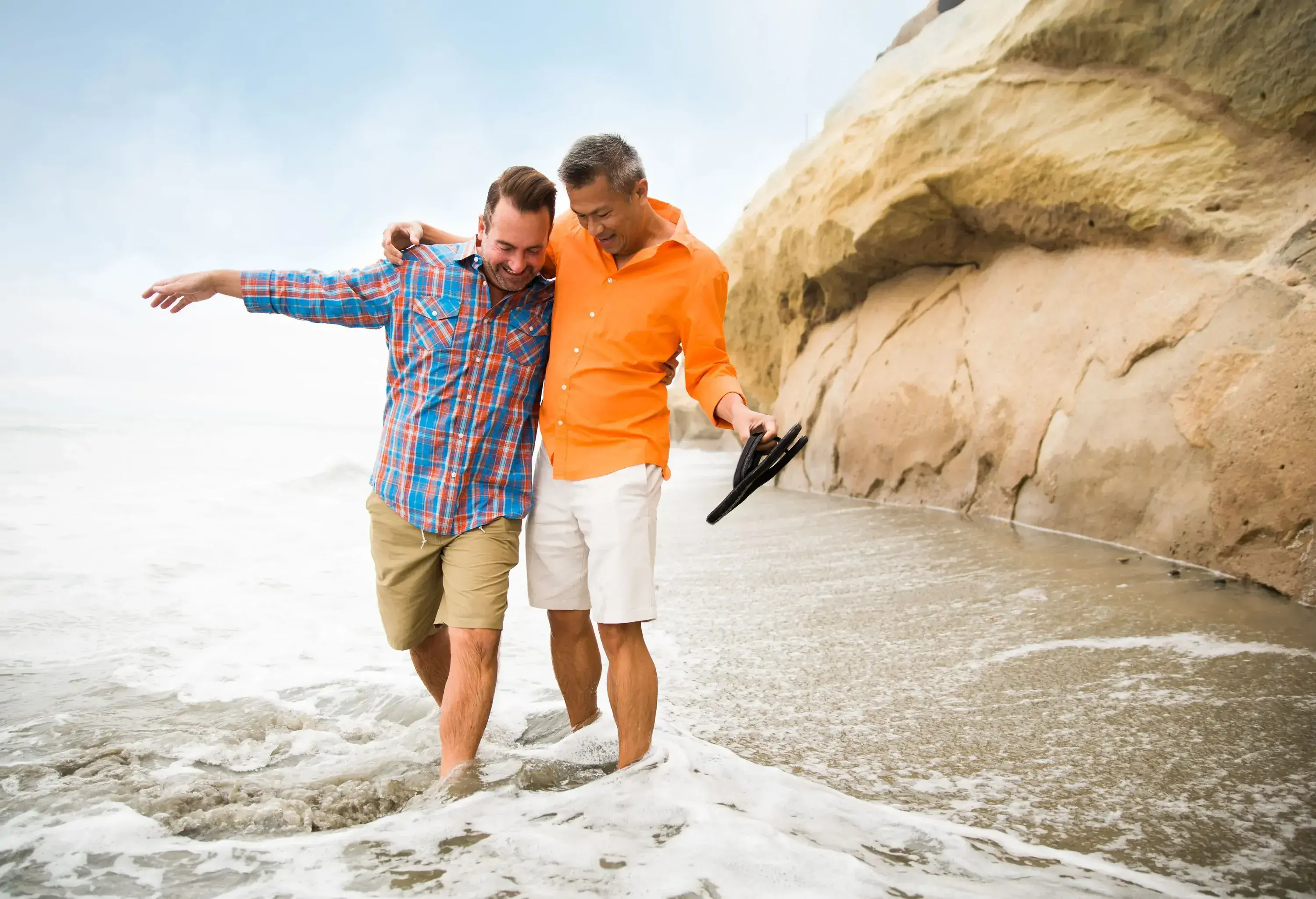 A smiling couple with their arms around each other stand in the shallow ocean surf on a beach along a rocky cliff face.
