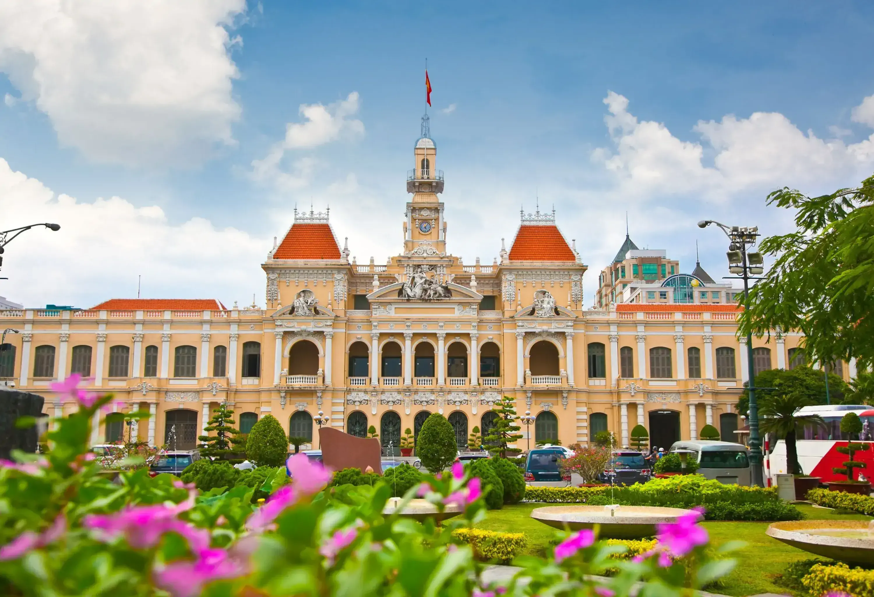 A building with pillared windows, orange roofs, and clock tower in front a park with curated gardens.