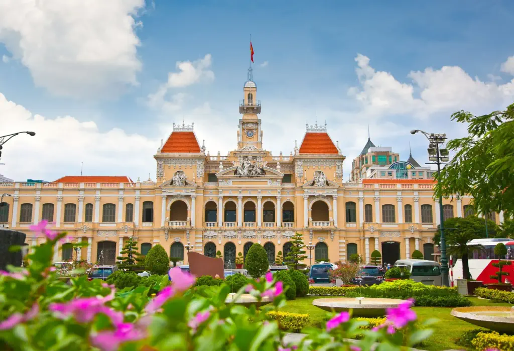 A building with pillared windows, orange roofs, and clock tower in front a park with curated gardens.