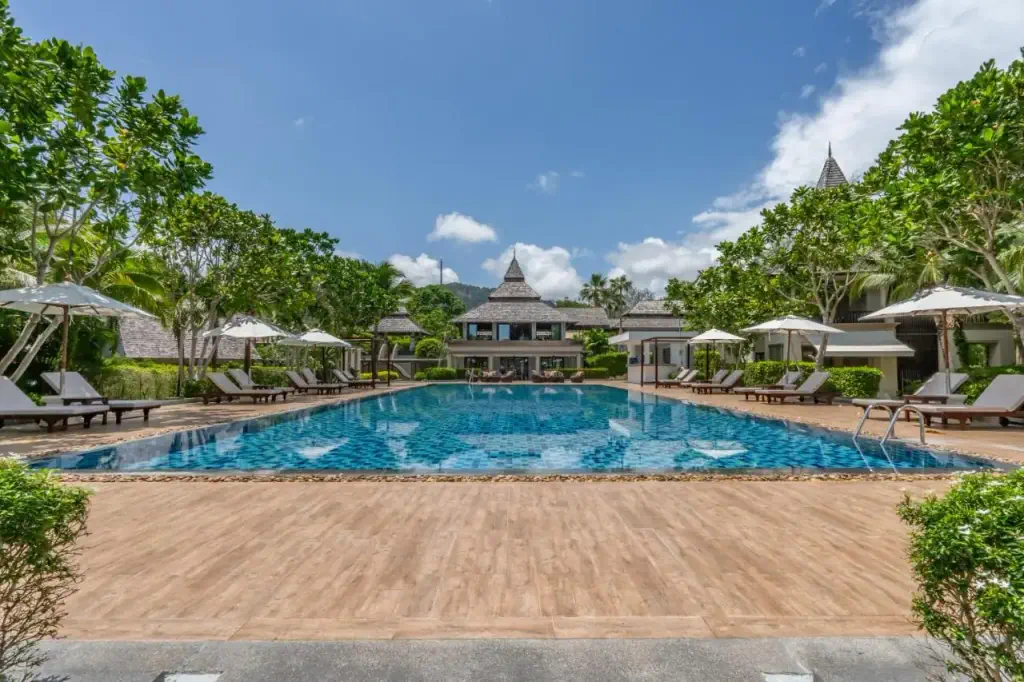 Luxurious resort pool surrounded by trees and sun loungers under white umbrellas. Blue sky and a traditional Thai-style building in the background convey a serene, tropical atmosphere.
