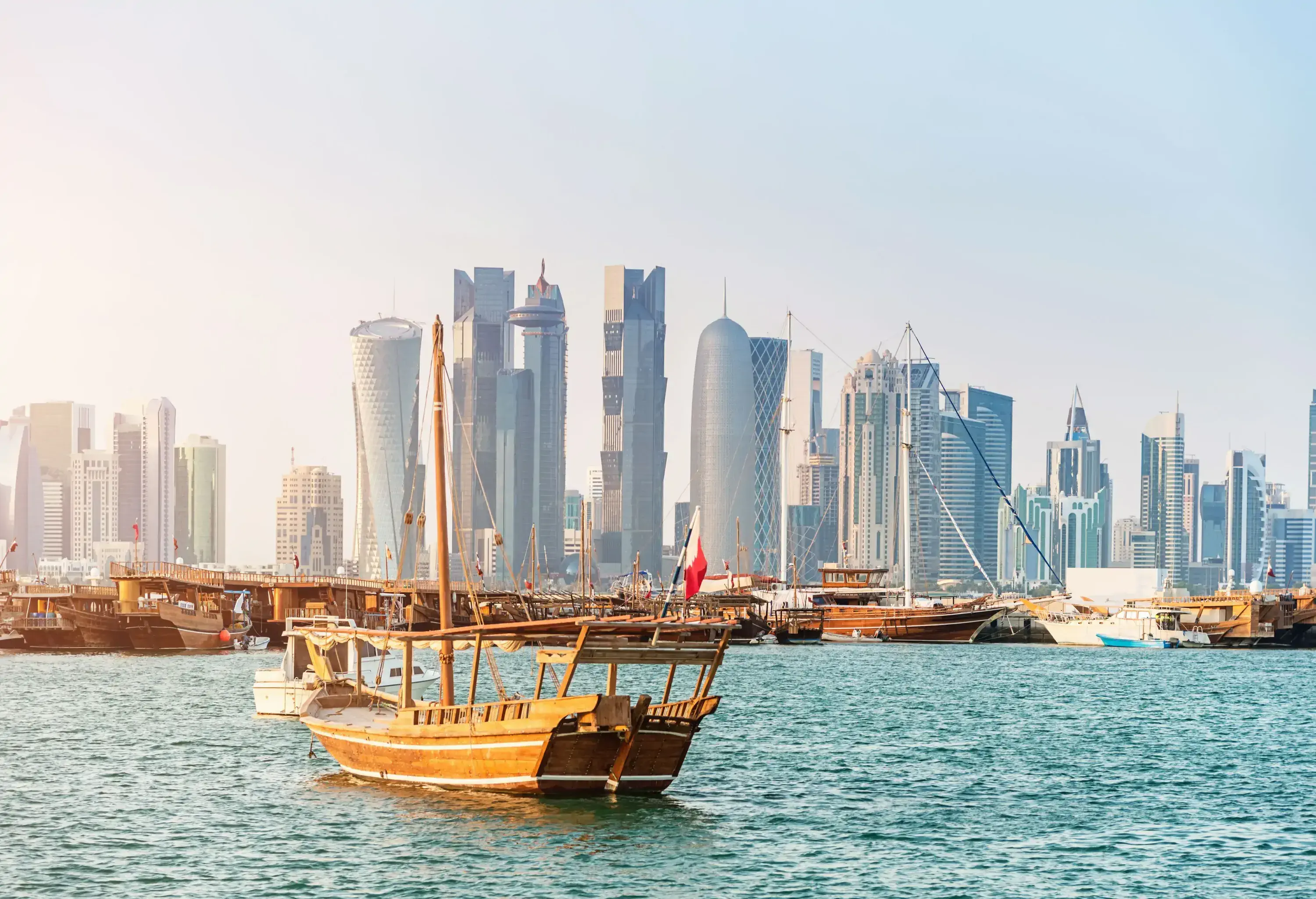 A wooden sailboat rocking in the ocean with striking skyscrapers lining the shore.
